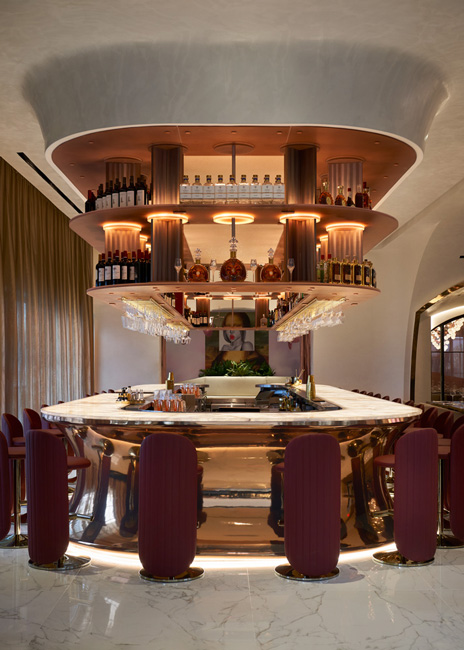 The horseshoe-shaped bar pays homage to the Hôtel Plaza Athénée in Paris. The translucent acrylic bottle rack stands above the bar, affectionately dubbed the “liquor chandelier.” It was custom crafted in Colorado and features rose-colored shelves illuminated from within while discreetly supporting more than 5,000 pounds of liquor bottles.